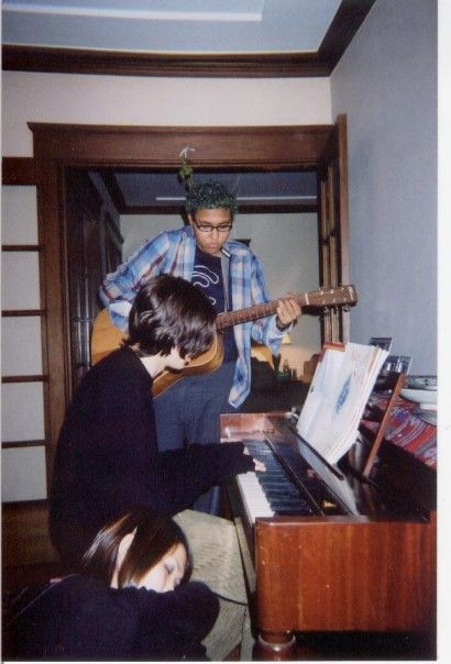 Three teenagers gathered around a piano. Bear, a light skinned man of color with blue curly hair and thick black-rimmed glasses stands playing a guitar. He is wearing a plaid print shirt, and a Belle & Sebestian t-shirt visible underneath. In front of Bear, Jordaan Mason sits at piano. Their face is obscured by their medium length black hair. Sitting beside Jordaan, Laura Jew sleeps with her head resting on the piano bench near Jordaan. The image is grainy - the texture of the matte print photograph in the image visible.