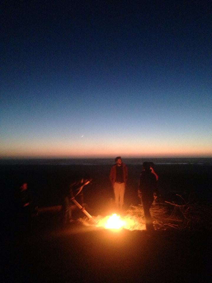 Bear stokes a beach bonfire. The ocean sunset is visible in the background, a faint sliver crescent Moon visible above the ocean horizon. In the foreground a group of people stand gathered around the warm glow of a campfire. All faces are obscured in the evening dusk light.
