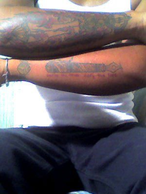 Image description: a grainy photo of Bear sitting on a chair holding his arm stacked so you can see the tattoos on his forearms: one is pen with writing which can't be made out in the photo below it. The other tattoo is a flaming sword.