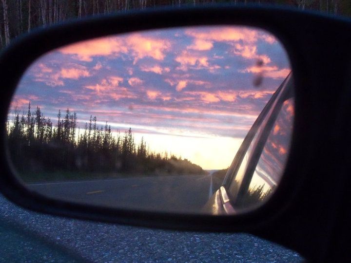 A photo of a car's driver side mirror with the sunrise sky and road in focus in the mirror. The clouds are lit up pink, orange, and purple. The bright yellow sunlit sky is visible below the cloud line above the horizon.